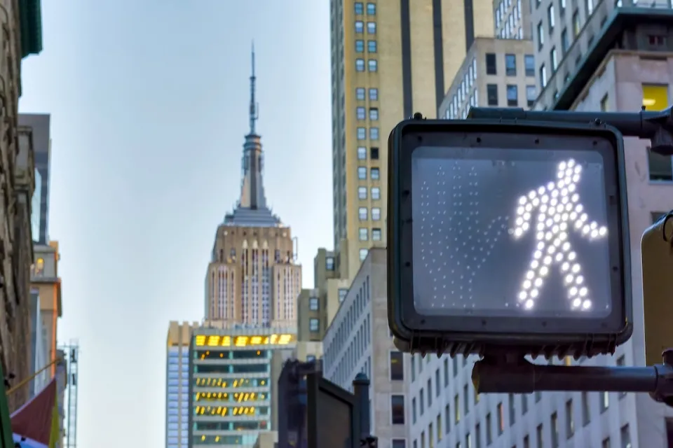 Close-up on pedestrian traffic signal with walk symbol lit up and Empire State Building spire visible in background.